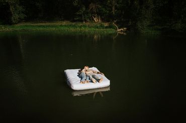 A couple lies barefoot on a floating mattress in a calm lake surrounded by trees.