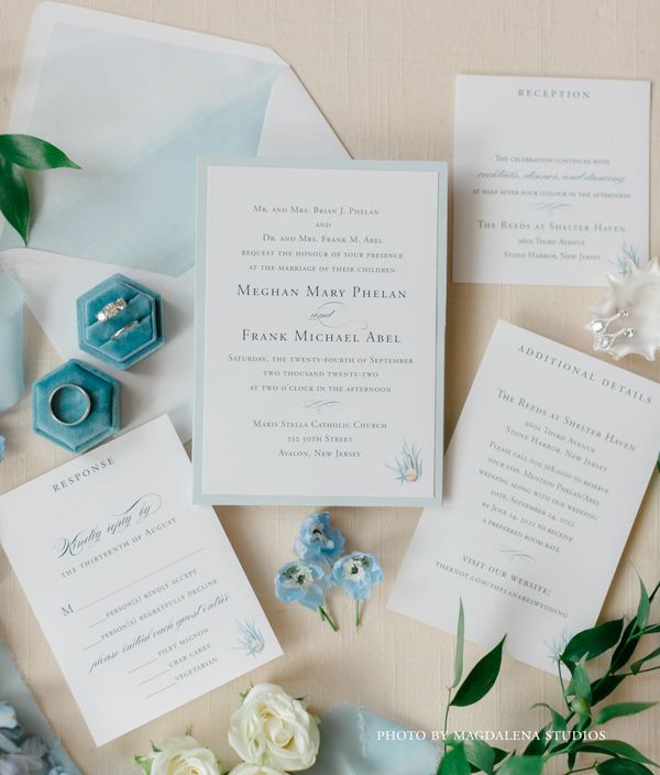 Elegant wedding invitation suite with rings, blue flowers, and greenery on a soft beige background.