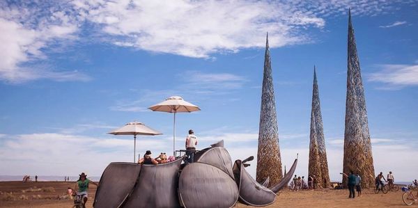 Large metal rhino sculpture with people and tall spire structures in desert under blue sky.
