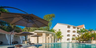 Sunny outdoor pool area with umbrellas and a multi-story hotel building.