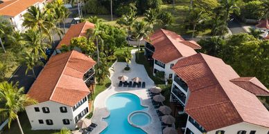Aerial view of resort buildings around a curved swimming pool with palm trees.