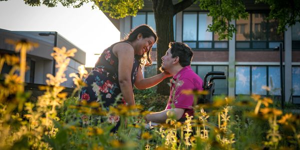 A woman in a flowered dress bends down to kiss her wheelchair bound fiancé in their engagement photo