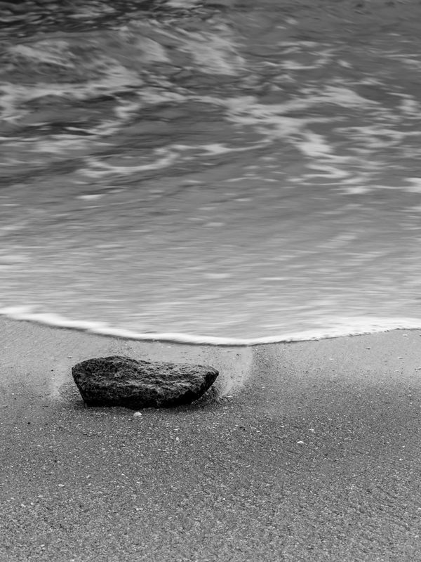 Black and white photo of a rock on wet sand with gentle waves.