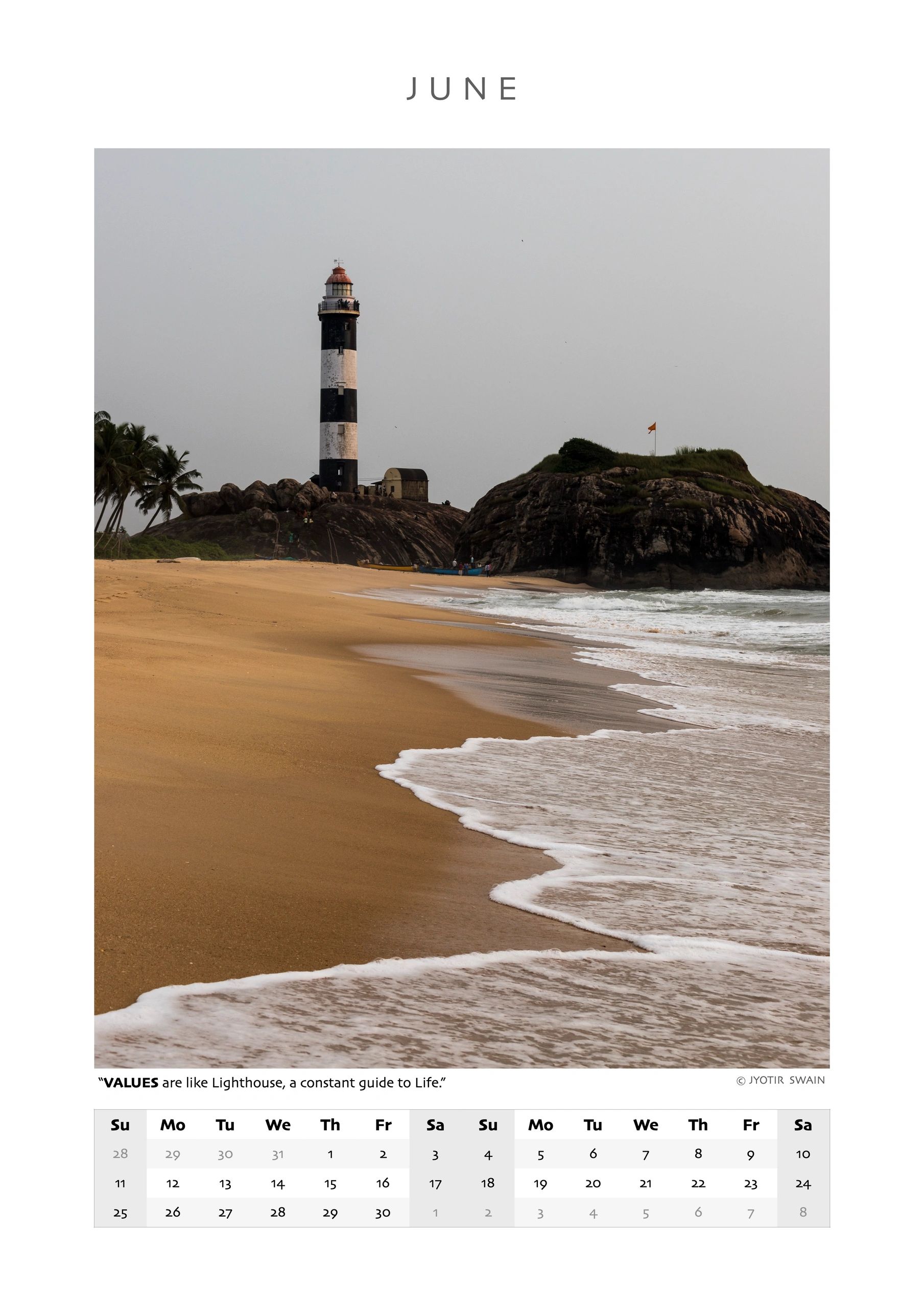 A black and white lighthouse on a rocky shore beside a calm beach in June.