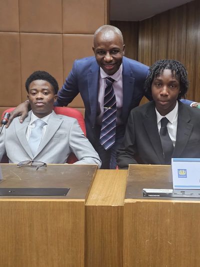 Three men dressed formally, two seated and one standing behind them, in a wooden-paneled room.