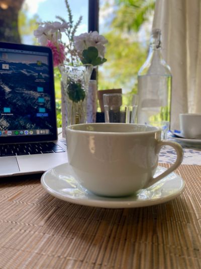White coffee cup on a saucer near a laptop and flowers indoors.