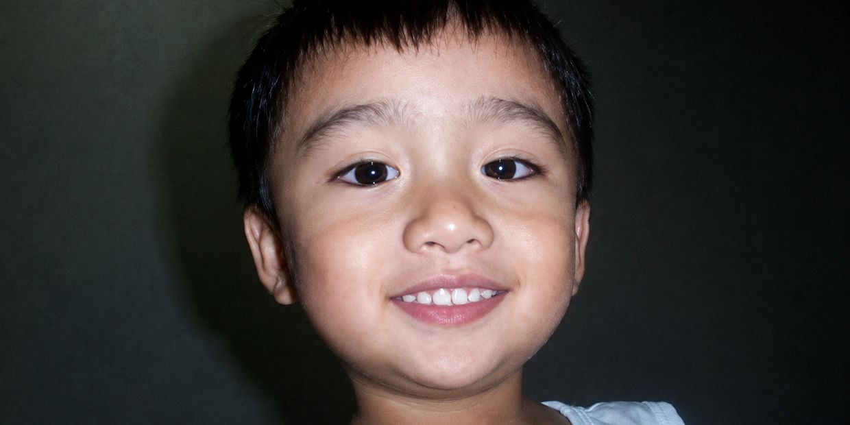 Close-up of a smiling young boy with dark hair against a dark background.