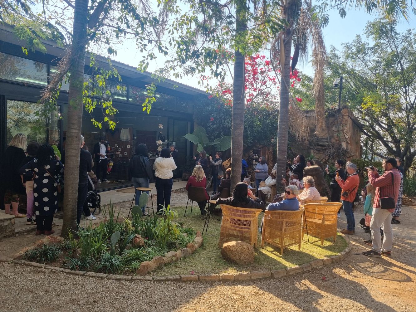 Outdoor gathering with people listening to a speaker near a garden and house.