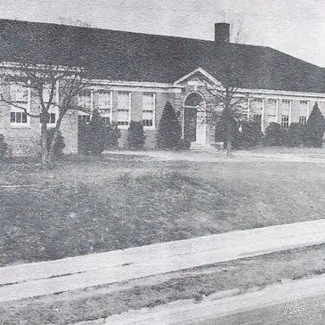 Black and white image of an old school building with trees and a sidewalk.