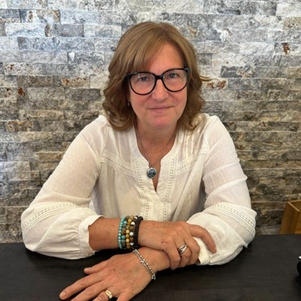 Woman in glasses and white blouse sits at a table with hands folded.