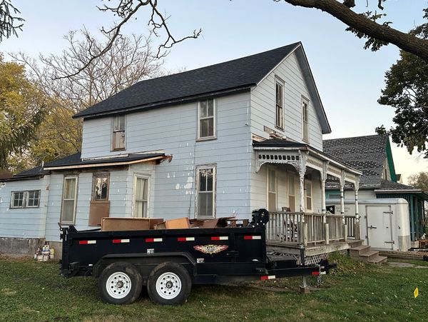Old house with peeling paint and a trailer full of debris in the yard.