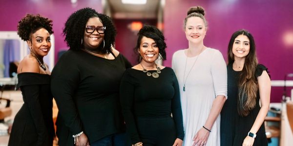 Five diverse women standing together smiling inside a room with purple walls.