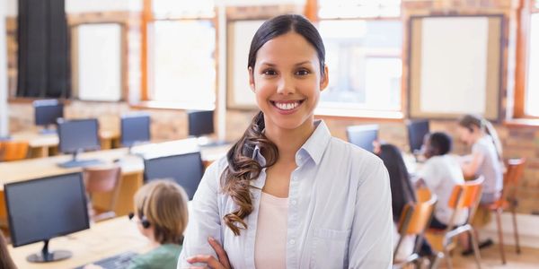 Smiling female teacher standing confidently in a classroom with students at computers.