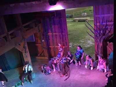 Actors perform a dramatic play on a rustic stage with purple lighting.