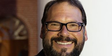Smiling man with glasses and beard against a blurred background.