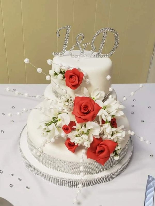 Elegant white wedding cake adorned with red roses and sparkling initials.