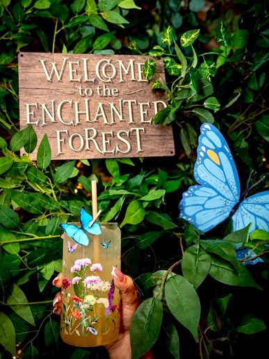 Welcome sign in a lush forest with butterflies and a floral drink.