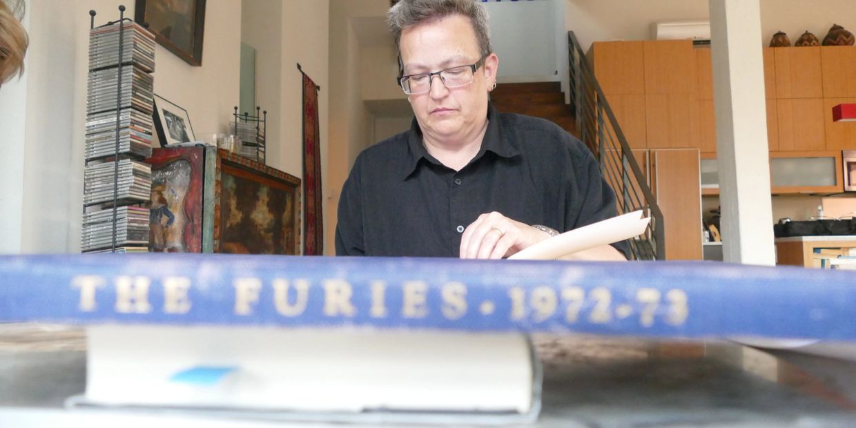 Jackie Rhodes sits at a table covered; in the fore in papers and books. In the foreground is a blue 