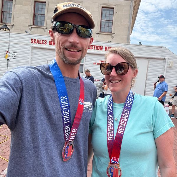 Two people wearing 'Remember Together' medals and sunglasses, smiling outdoors.