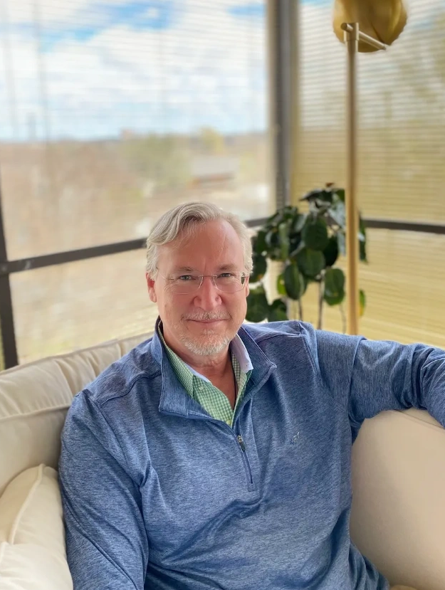 Man sitting comfortably on a beige couch in a sunlit room with blinds.