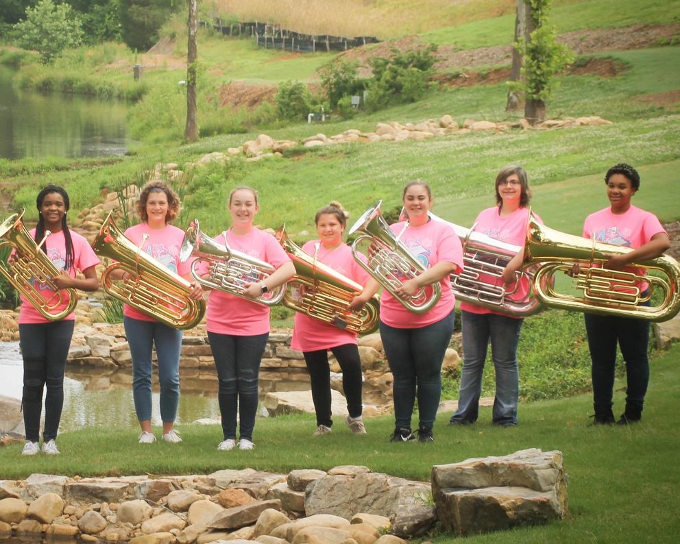Seven young women holding brass tubas, standing on grass near a pond.