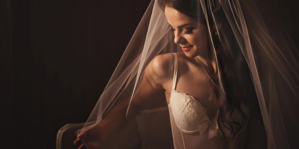A woman in white lingerie smiles softly under a sheer veil in warm lighting.
