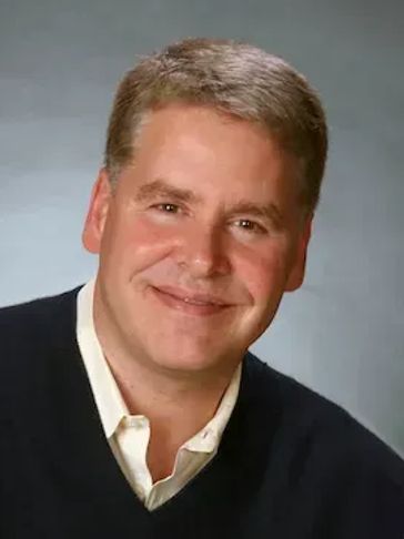 Smiling middle-aged man in a black sweater and white shirt against a gray background.