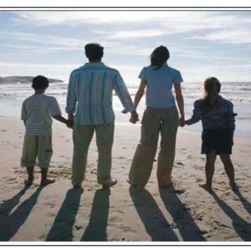 A family of four holding hands and facing the ocean on a sandy beach.