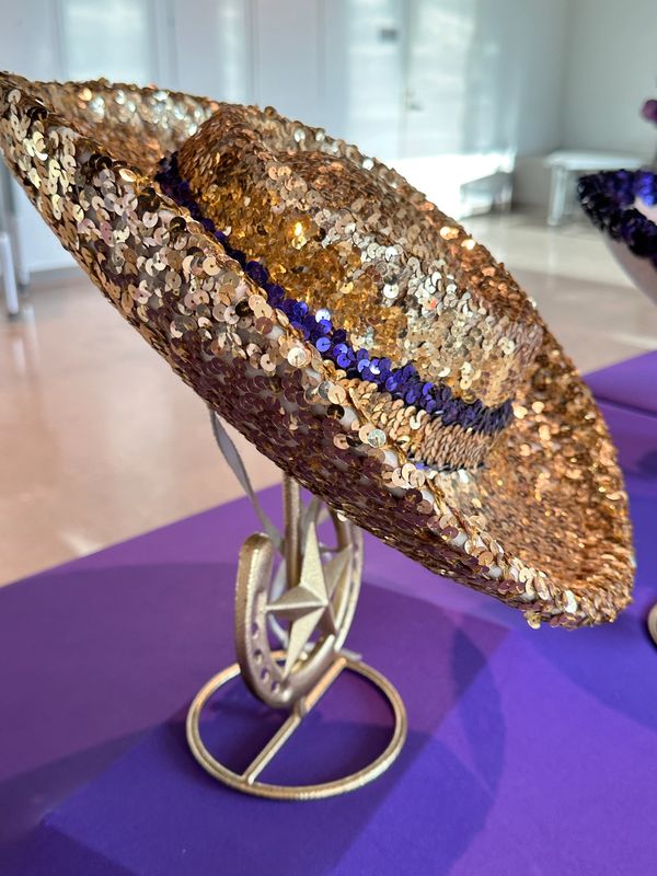 A shiny gold sequin hat with a blue band on a metal stand.