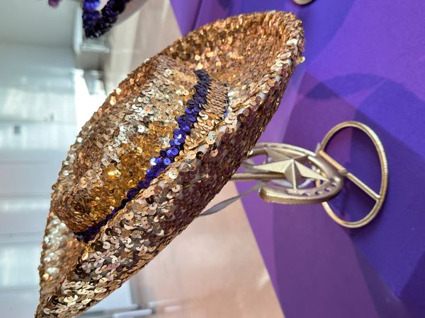 A shiny gold sequin hat with a blue band on a metal stand.