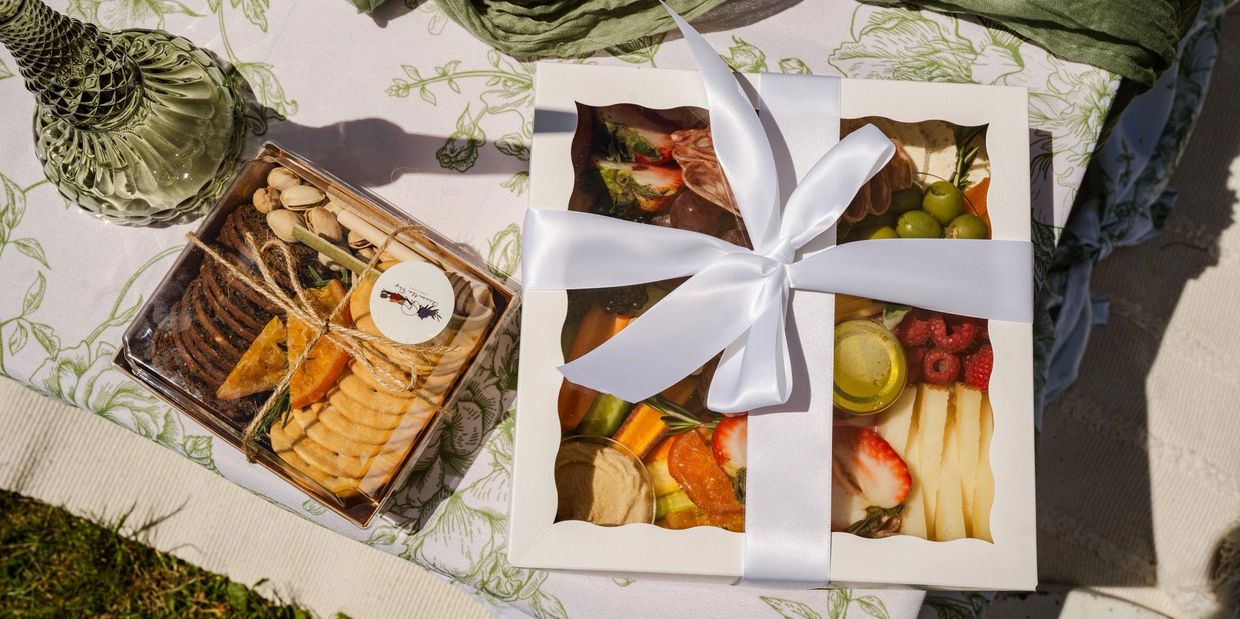 Two elegant gift boxes with assorted snacks and fruits on a floral tablecloth outdoors.