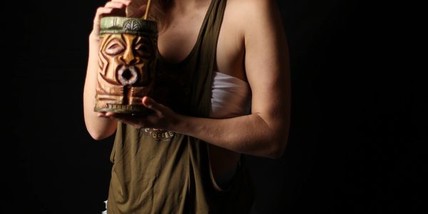 Person holding a tiki-style cup with a straw against a black background.