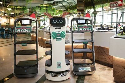 A cat-like delivery robot wearing reindeer antlers between two delivery robots wearing Santa hats.