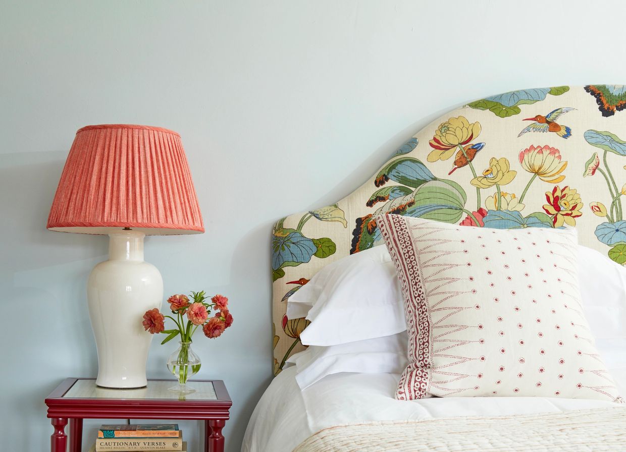A bedside table with a lamp and flowers next to a bed with a floral headboard.