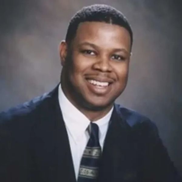 Smiling man in a suit and patterned tie against a dark backdrop.