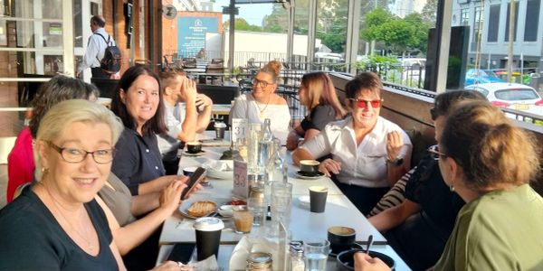 Group of women enjoying coffee and conversation at a long table in a bright cafe.