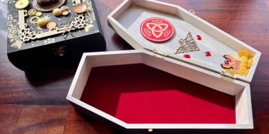 Open wooden coffin-shaped box and decorative black box on wooden floor.