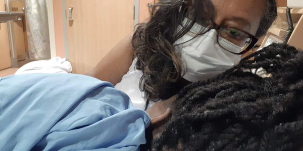 A person wearing a mask comforts another lying down in a hospital room.