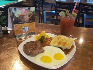 Steak and eggs breakfast on a table with a bloody mary on a plate