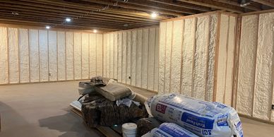 A basement under renovation with spray foam insulation on walls and construction materials on the floor.
