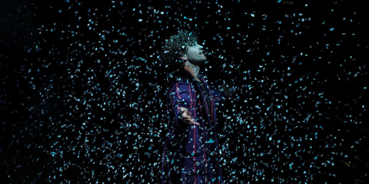 Man joyfully standing amidst falling blue confetti against a dark background.