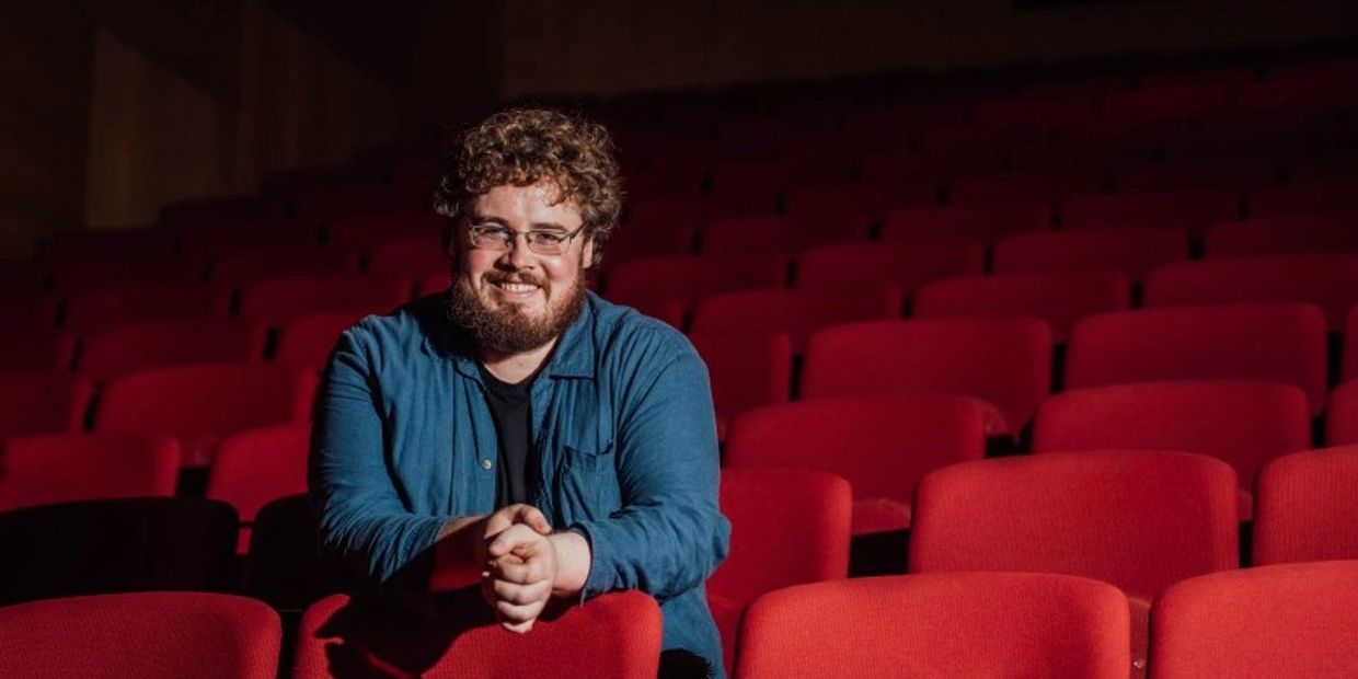 A smiling man with curly hair and glasses sits among red theater seats.