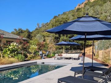 Sunny outdoor pool area with loungers and umbrellas against a hillside.
