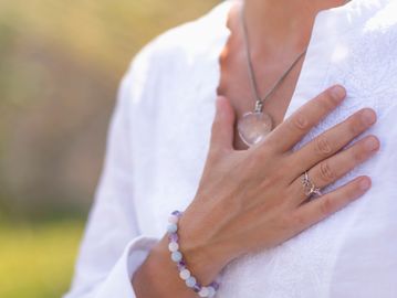 Person placing hand on chest wearing a white shirt and jewelry.