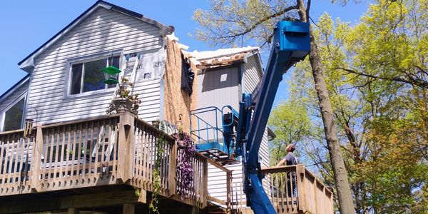 A man using a blue boom lift to work on a house exterior under bright blue skies.