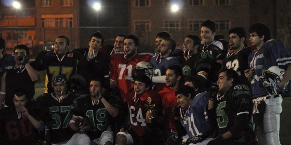A group of young athletes posing with medals and a trophy on a football field at night.