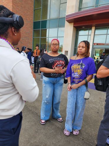 Two women stand outside a building, being interviewed by a person with headphones.