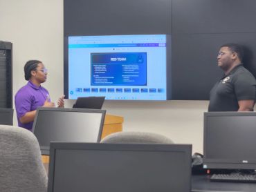 Two men discussing a presentation in a classroom with a slide about the Red Team.