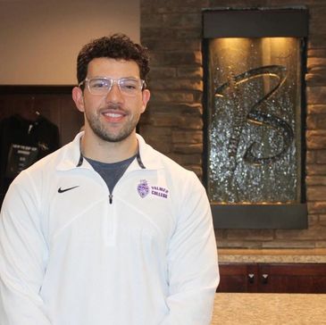 A man in a white Nike jacket standing indoors with a stone wall and decorative panel behind him.