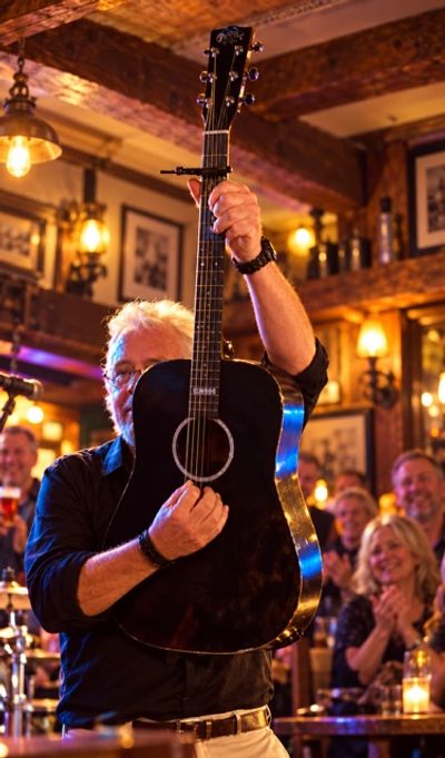 Man holding a black acoustic guitar in a lively pub.